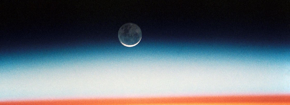 The Earth's atmosphere seen edge-on at sunrise from orbit, a thin band of blue and orange beneath a crescent moon.